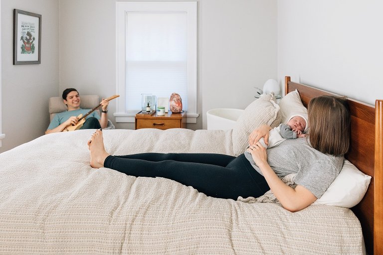 Mom holds baby on the bed while Dad plays guitar in the background.