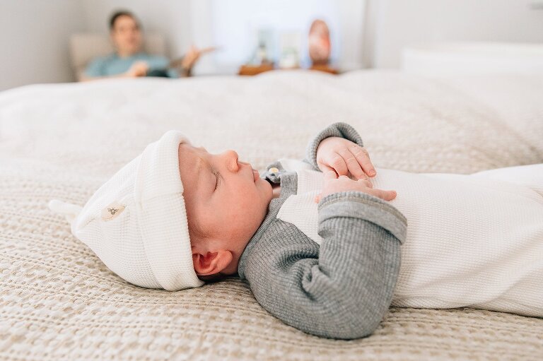 Newborn lies on the bed. Dad is in the background playing the guitar. 