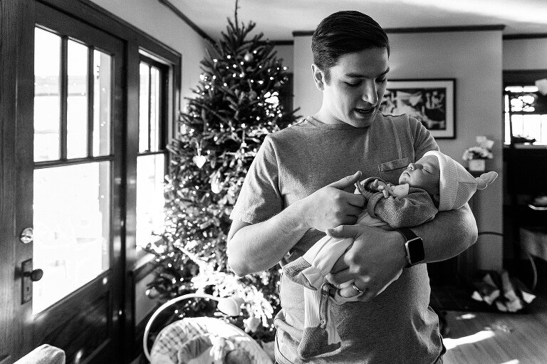 Black and white. Dad holds newborn and reaches for his fingers. Christmas tree in background. 
