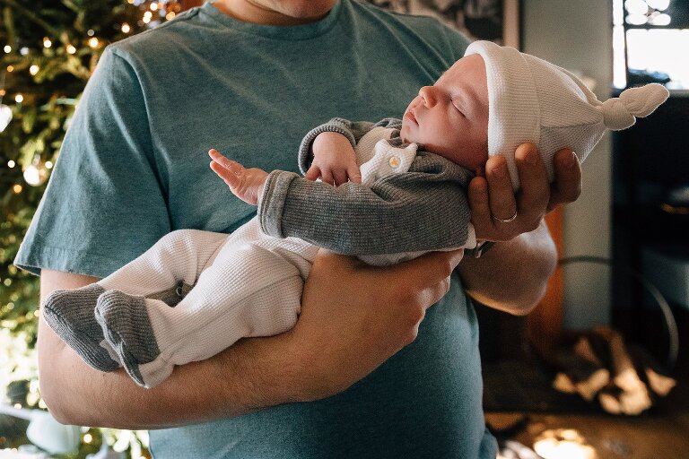 Dad holds baby, Christmas tree in background. 