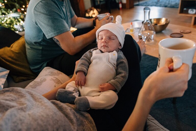 Mom holds newborn in her lap and cup of coffee in hand; Dad sits next to her holding coffee mug. 