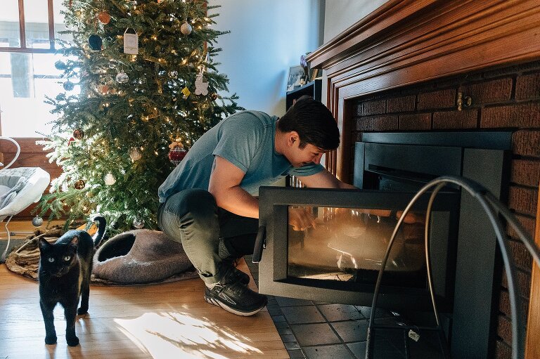 Man stokes fire in fireplace while black cat stands behind him. 