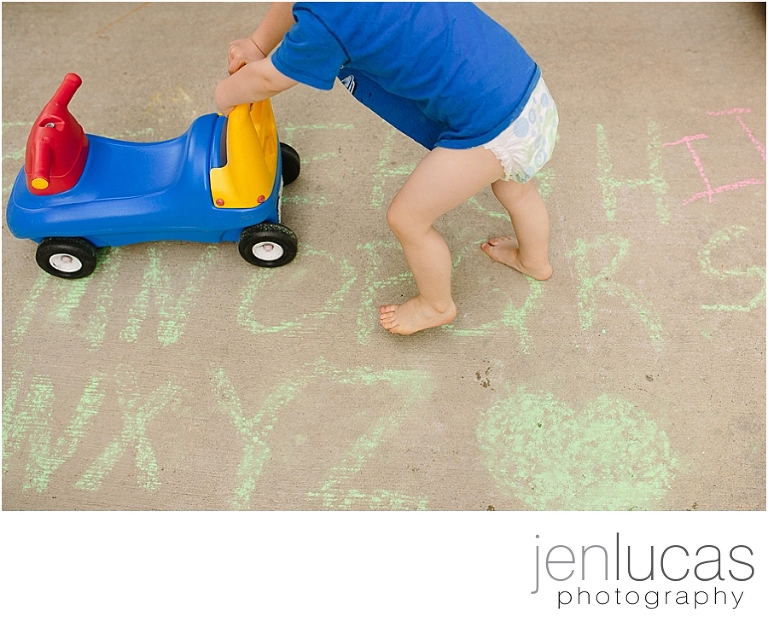 A toddler pushes a toy across the driveway that is full of chalk writing of the alphabet
