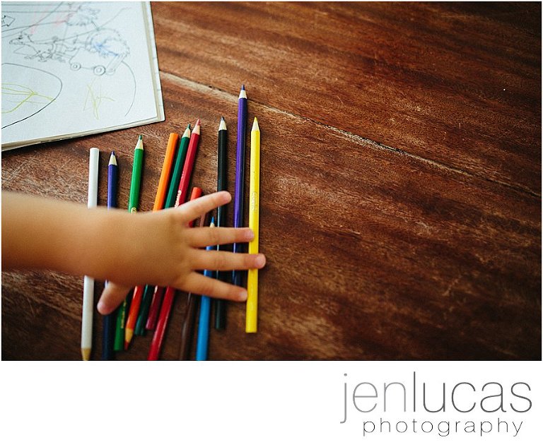 A child's hand reaches across a line of colored pencils. A coloring book is in the upper left corner of hte image. 