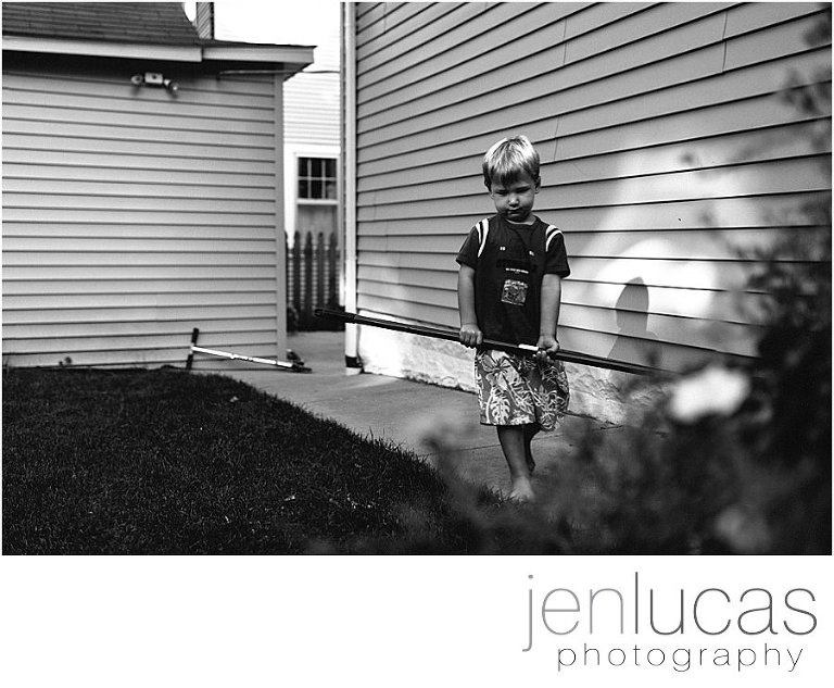 A preschool boys carries a shovel in the backyard. He steps into a pocket of light the produces his shadow on the house. 