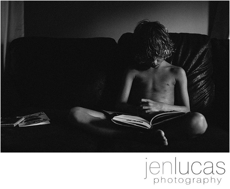 Black and white. A young boy sits on a dark couch and reads a book. There is subtle window light on the frame. 