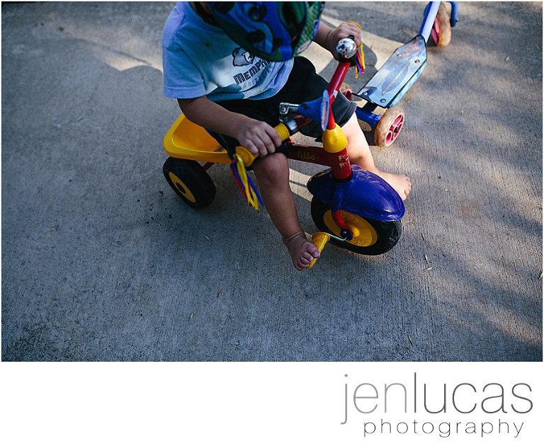 top down view. Child rides a tricycle in shade and pockets of sunlight. He is barefoot. 