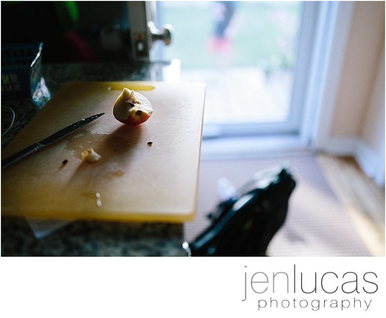 A cut apple on a yellow cutting board, backpack on the floor, a child outside in the background 