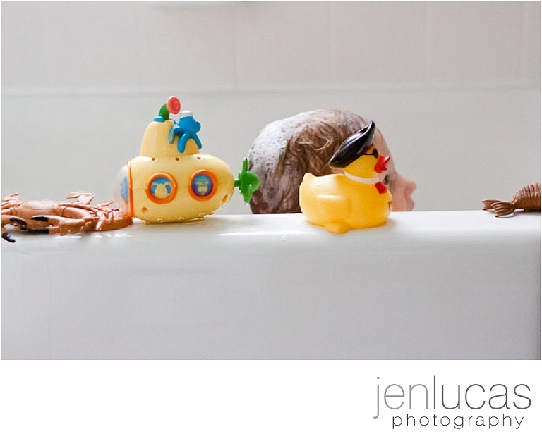 Bath toys line the edge of the bath tub while a small boys' face mimics the face of a rubber duck