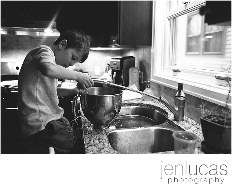 A young boy sits on a step stool while he tries to wash out a mixing bowl with the faucet extension. Water splashes onto his pants. 