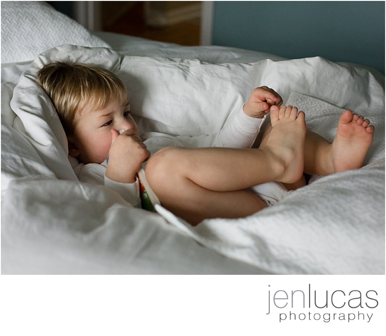 A young boys sits in a white bed, reaching for his toes. 