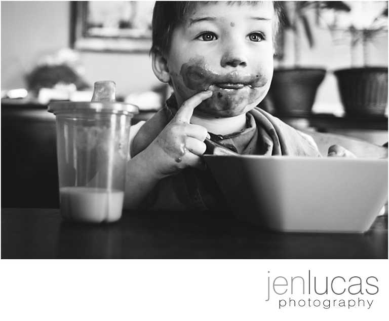 Black and white. A toddler has an extremely messy face as he eats a bowl of tomato soup. There is a sippy cup of a small amount of milk next to him. 