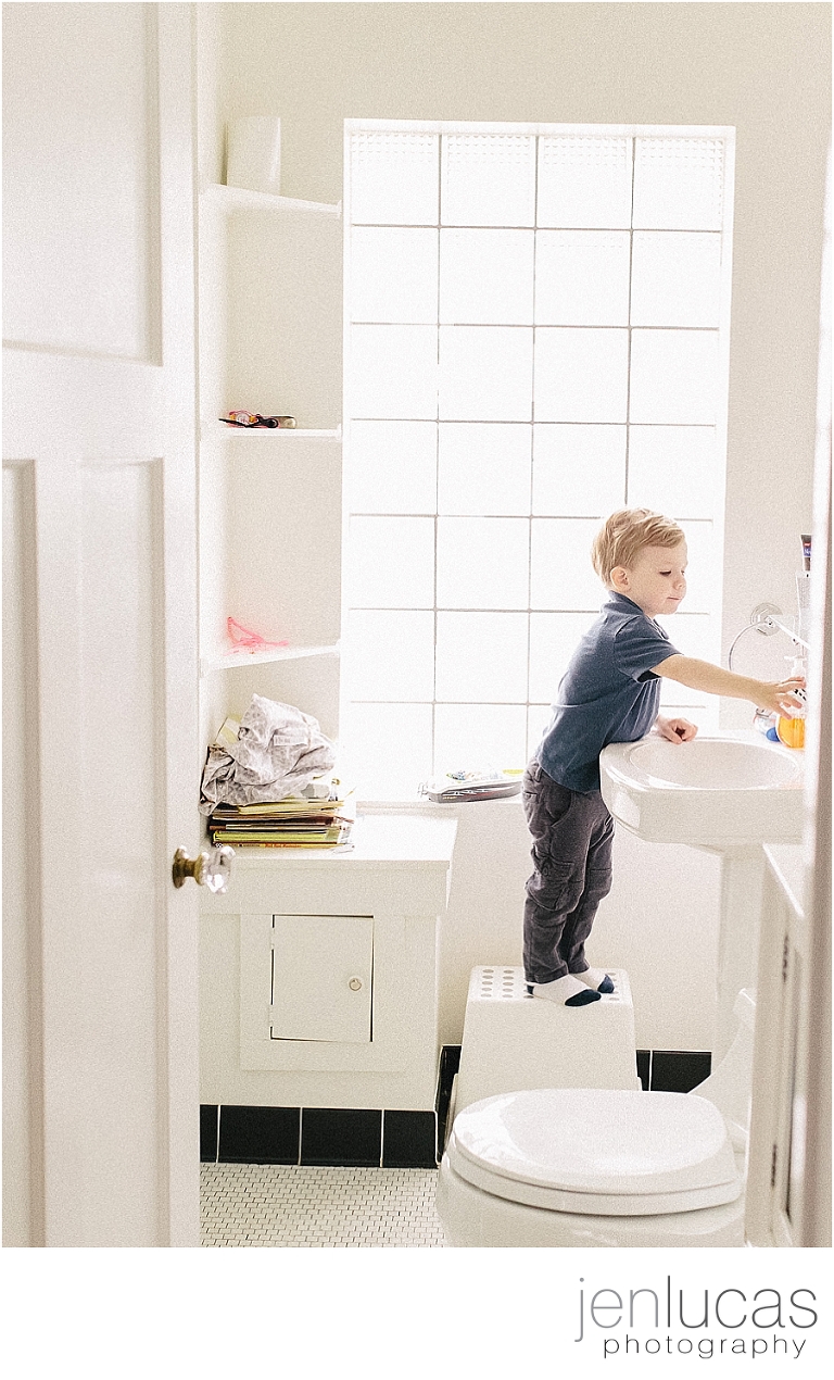 Boy stands in on step-stool in a white bathroom as he reaches for a yellow soap container on the sink.