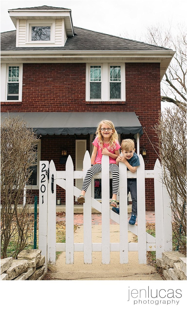 A boy and girl climb the white fence gate that surrounds their home. 