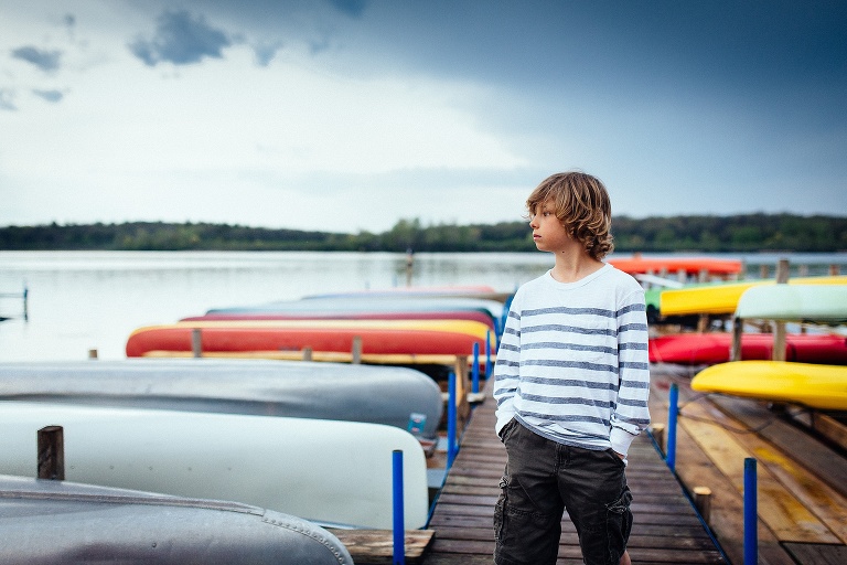 Boy standing on a dock next to many canoes. Dark cloudy sky.