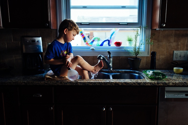 Young boy sitting on the kitchen counter with his feet in the sink washing off his muddy feet.