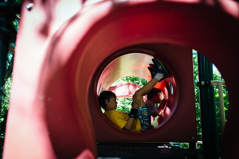 Two in a red tunnel at a playground. Boy laying down with his feet in the air while the other looks out a circle window.