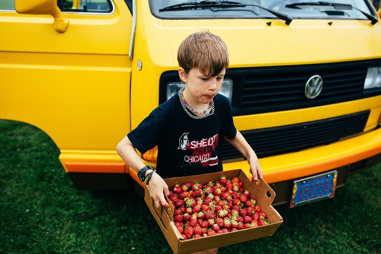 Young boy stands in front of a old bright yellow van holding a box of fresh strawberries.