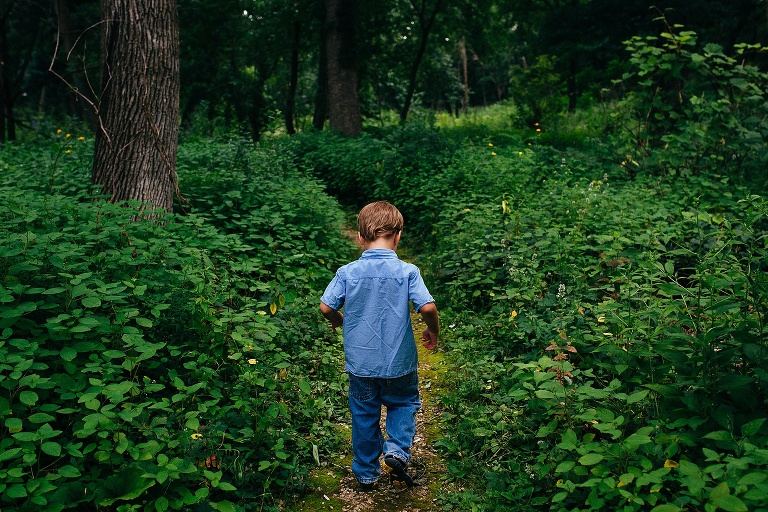 Young boy walks down a narrow path outside next to green bushes and tall trees.