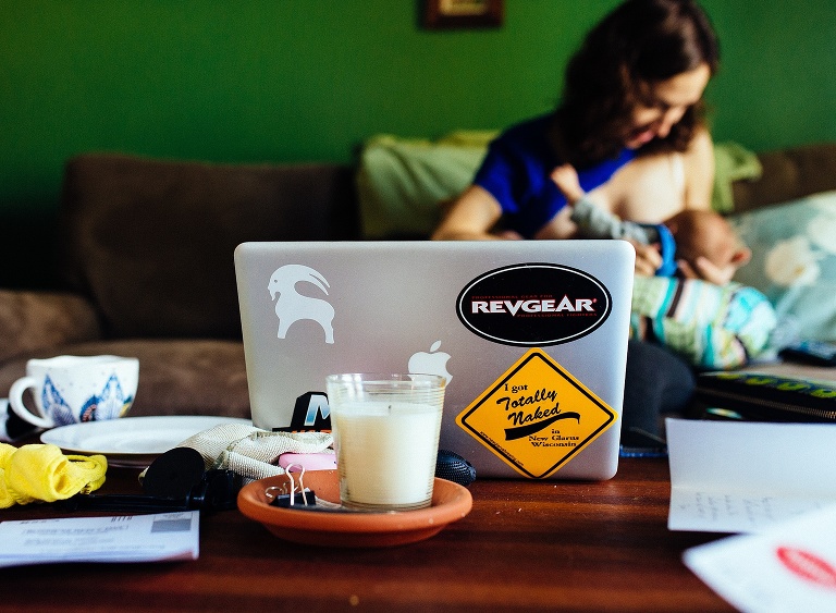 Laptop laid out on messy coffee table as mother breastfeeds her newborn baby boy.