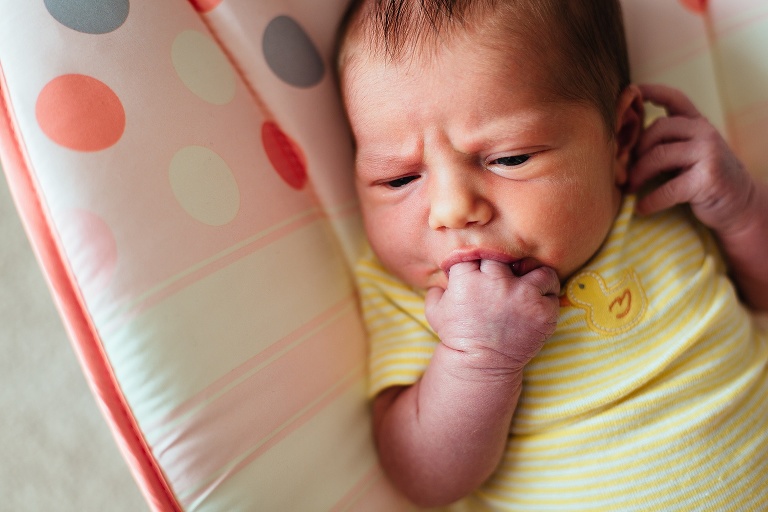 Newborn baby wearing a yellow duck onesie sucking on her fingers.