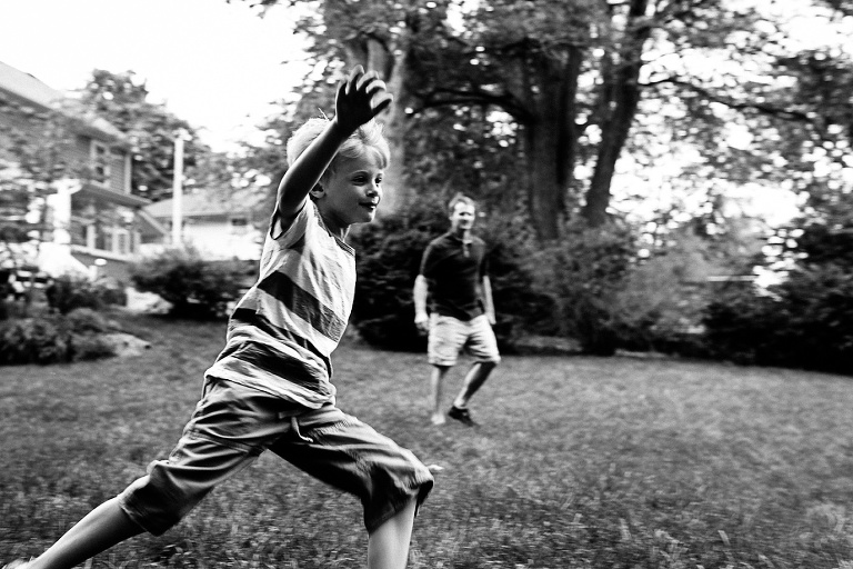 Black and white image. Young boy running in his backyard, Father standing behind him.