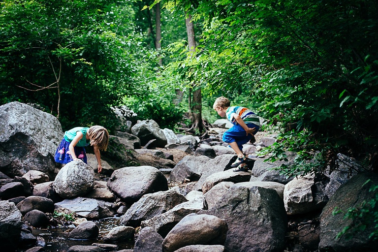 Brother and sister standing under many green trees climb on rocks in a small creek.