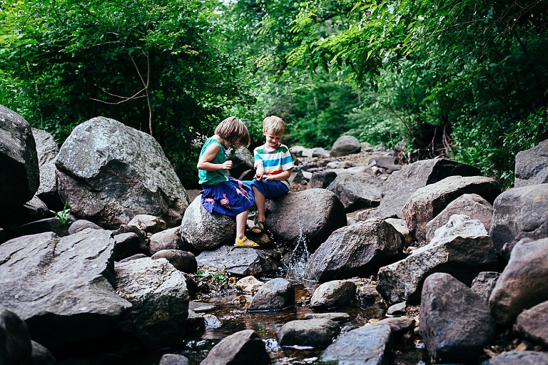 Brother and sister sit under the green trees sitting on the rocks in a small creek.