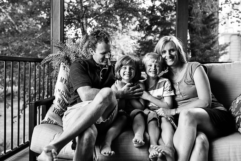 Black and white. Family of four sitting together on a couch outside on their back porch.