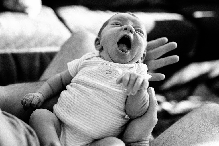 Black and white. Father holds yawning newborn baby over his lap.