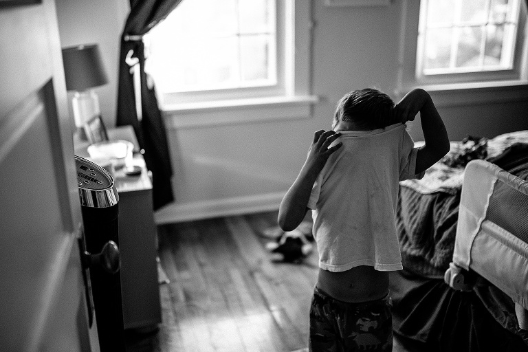 Black and white. Boy standing in his bedroom trying to pull his t-shirt over his head.