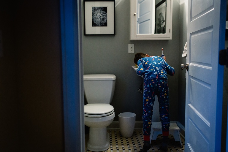 Boy wearing pajamas stands over sink in bathroom brushing his teeth early in the morning.
