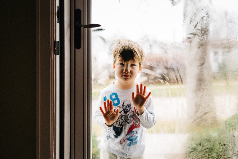 Young boy stands outside with face and hands against frosty cold glass door.