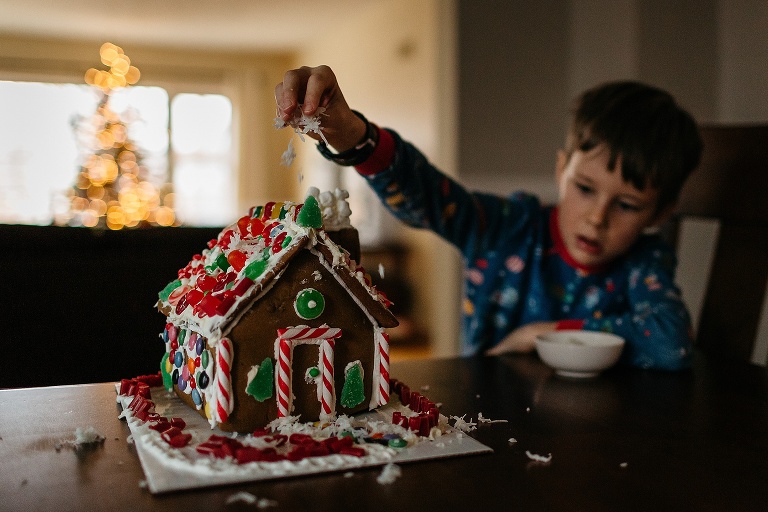Young boy sits at dinning table sprinkling fake snow on a gingerbread house. white lights from christmas tree shines in the background.