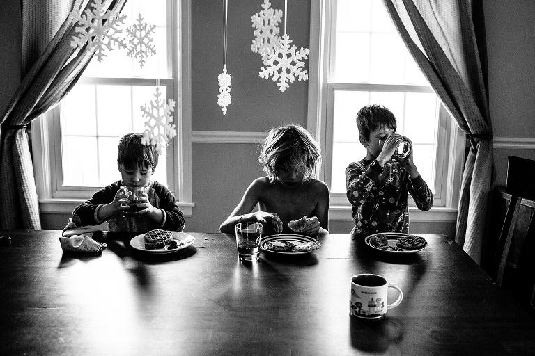 Black and white. Three siblings sit together at dinner table eating breakfast, paper snowflakes hang above them.
