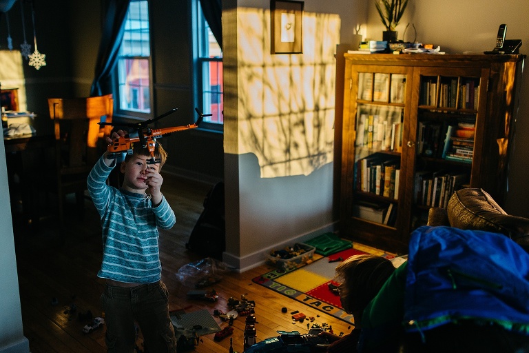 Young boy plays in living room with legos and holds toy helicopter in the air. windows shine light and shadows of bare trees onto the wall
