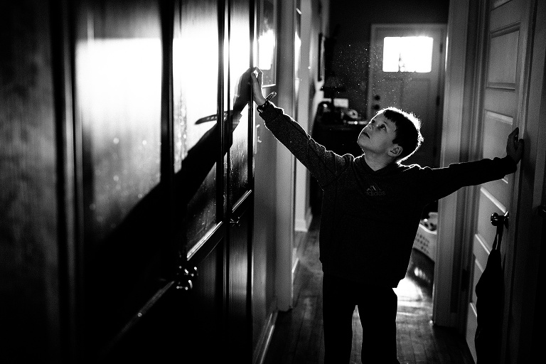 Black and white. Boy walks down hallway with both arms on the walls. Sun shining through the door window showing all the dust in the air.