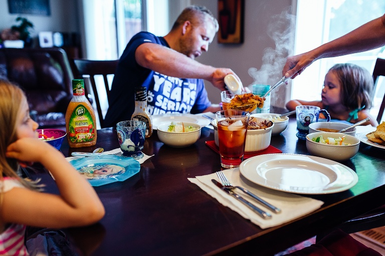 Family of four sits down to dinner at table. Steam rises from hot pasta as the mother lifts some pasta out of the container. Dad pours dressing on the salad and girls patiently wait. 