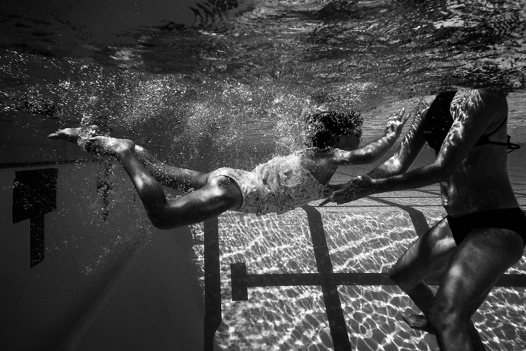 Black and white. Mom standing in the pool holds her hands out to catch her daughter jumping into the pool.