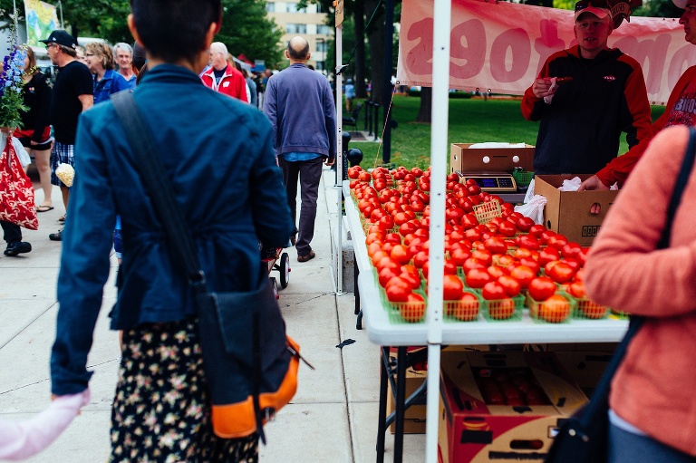 Mom holding her daughters hand walks through a farmers market standing next to a vendor selling bright red tomatoes.