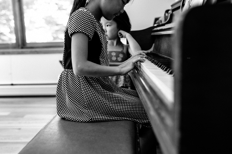 Black and white. Young girl sits and plays the piano while here sister stands next to her.