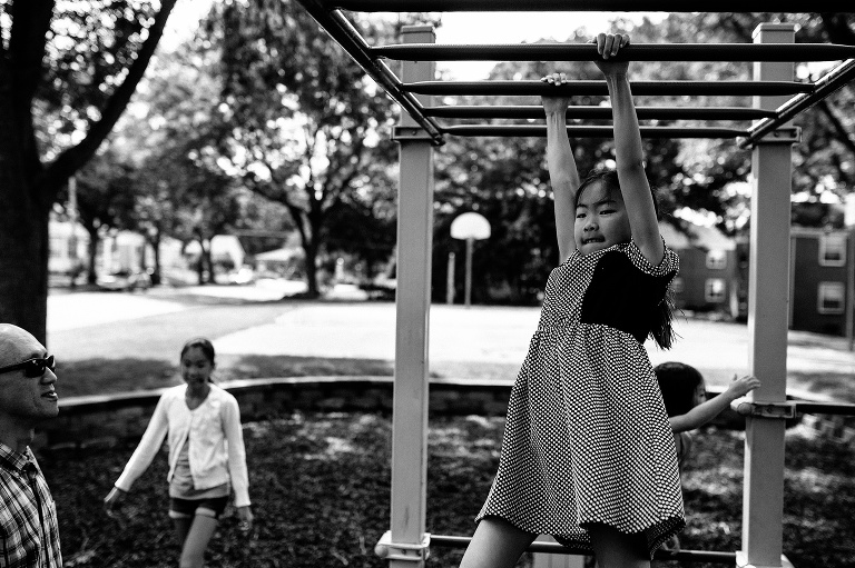 Black and white. Young girl swings across the monkey bars at a park.