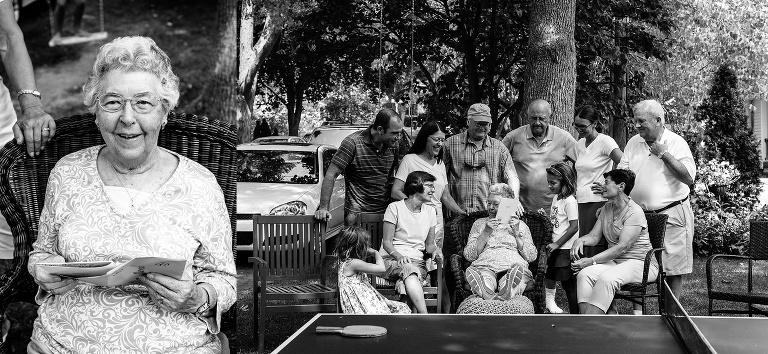 Black and white. Family stands around their 90 year old mother as she reads her birthday cards.