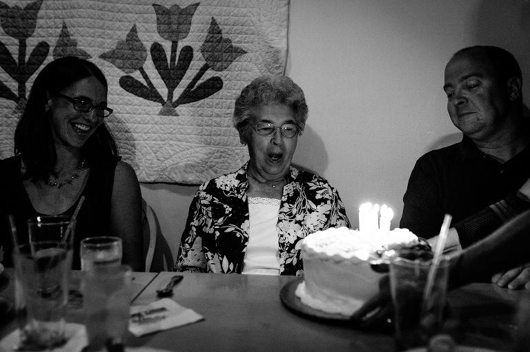 Black and white. Family sits around mother blowing out candles on her cake celebrating her 90th birthday.