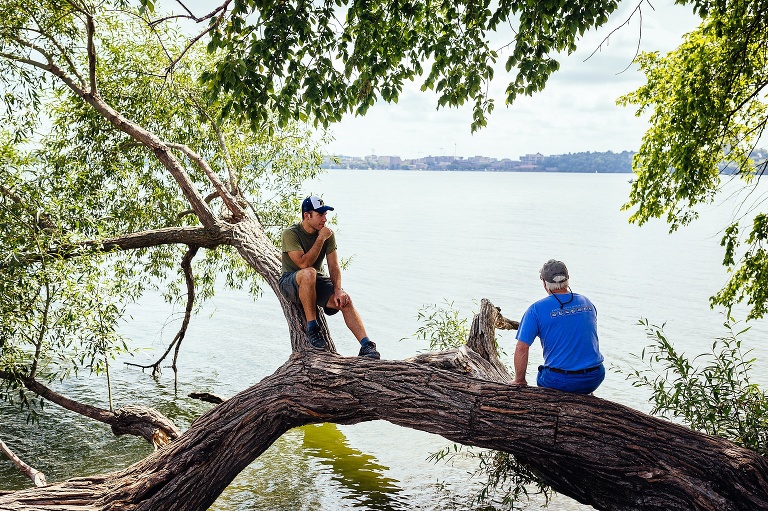 Father and grandfather sit on a fallen tree resting over the edge of a lake.