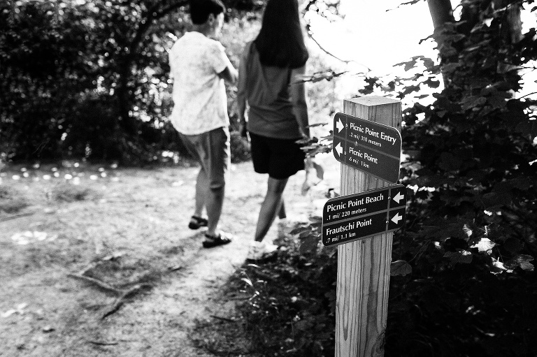 Black and white. Two women walk outside on a path next to a pole with directions of paths.