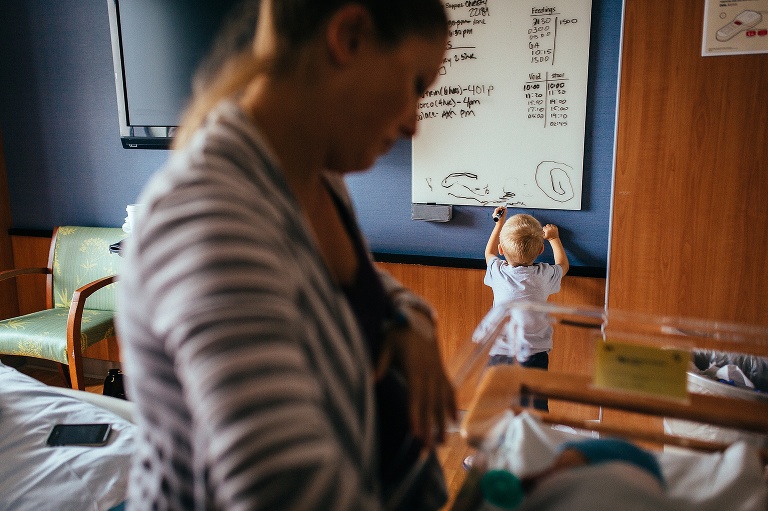 Mom standing in the hospital room looking at sleeping baby. Young boy standing behind her draws on the dry erase board on the wall.