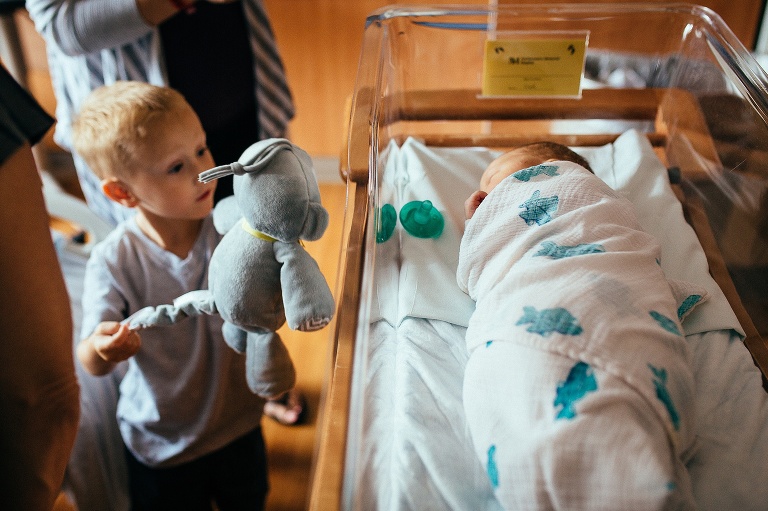 Young boy holds up a stuffed animal to his new baby brother.