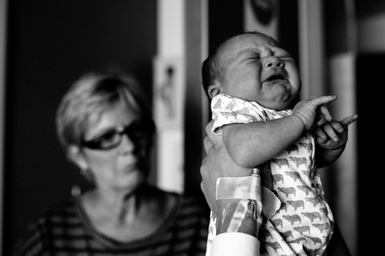 Black and white. Mom holds up crying newborn baby. grandma sitting behind them.