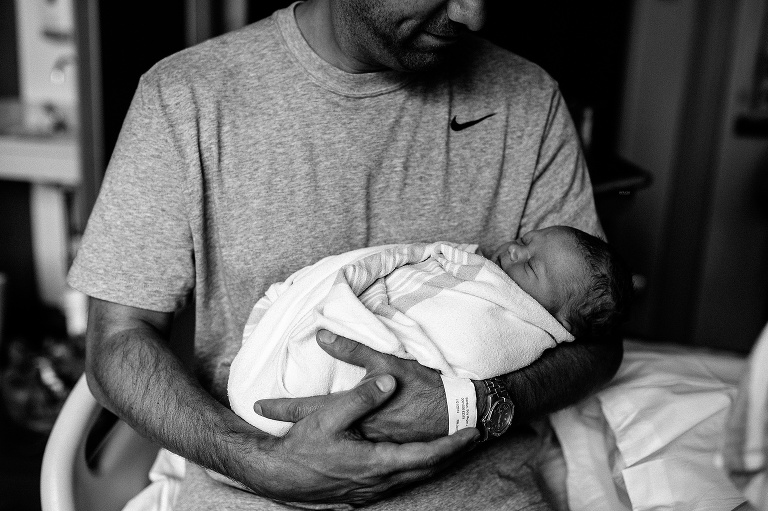 Black and white image. Father sitting in the hospital holding his newborn baby son.
